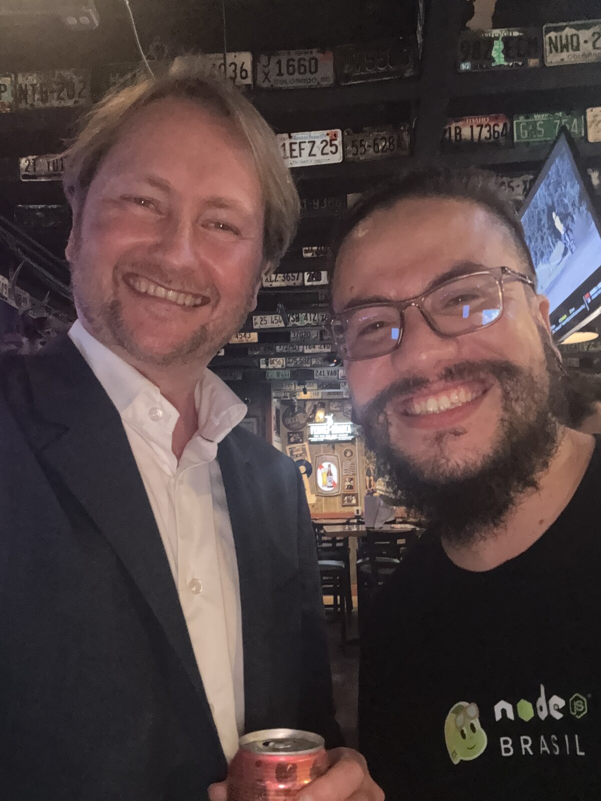 Matt Biilmann in a dark blazer and white shirt holding a can, next to Erick Wendel in a node.js Brasil t-shirt and glasses, both grinning, under a bar wall and ceiling covered edge to edge in old license plates.